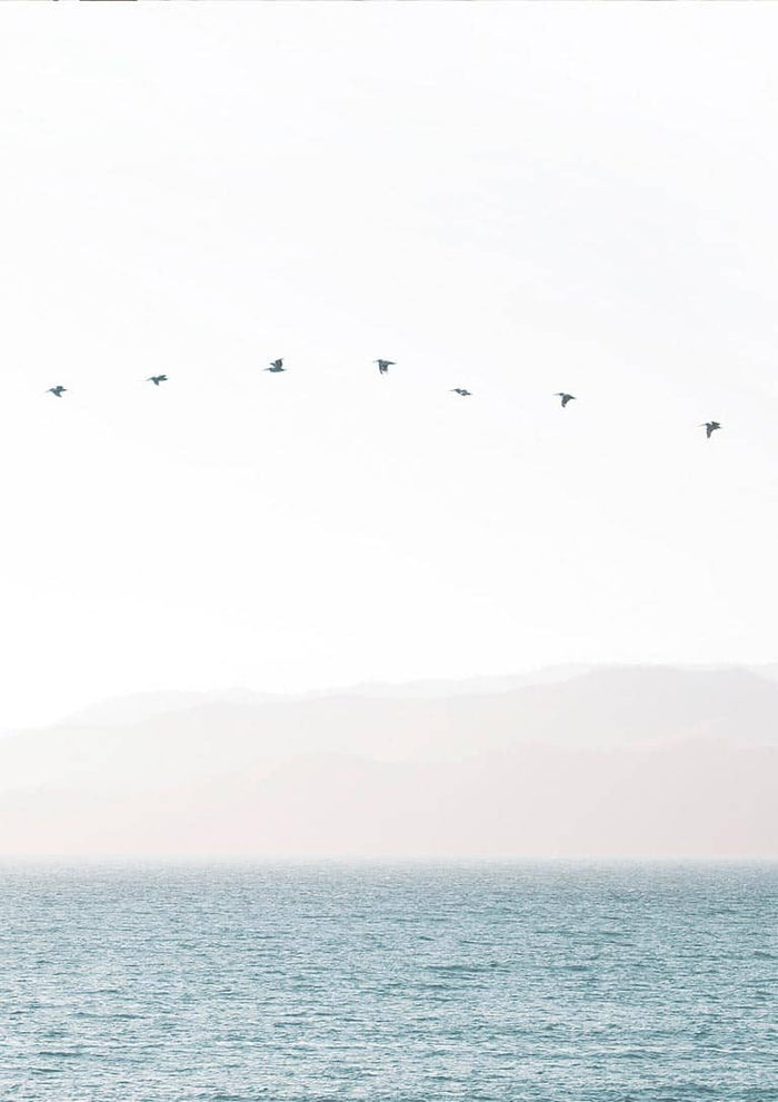 Minimalist artwork of birds flying in a straight line over calm water with soft mountains in the background.
