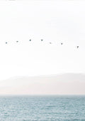 Minimalist artwork of birds flying in a straight line over calm water with soft mountains in the background.