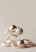 Three elegant glass Christmas balls and a cream ribbon displayed in and around a glass on a neutral background.