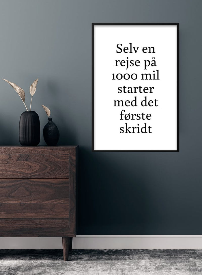 Det første skridt poster hanging on a dark wall above a wooden dresser with vase and dried grasses.