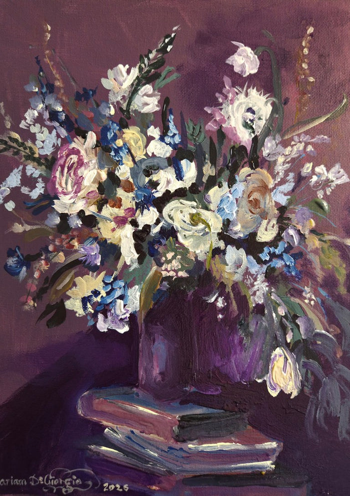 February arrangement by Mariam De Giorgio — a bouquet of cream roses and blue blossom in a plum vase on stacked books against a deep purple ground.