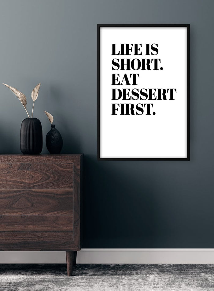 Minimalist "Life is Short. Eat Dessert First." poster on a dark wall above a wooden dresser with decor.
