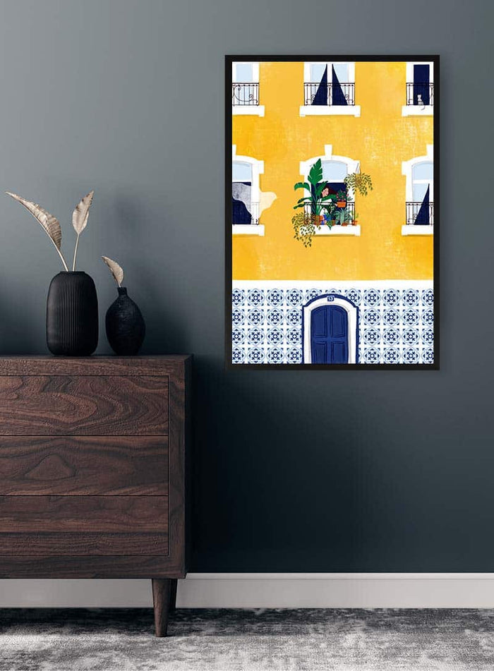 Framed artwork of a yellow Lisbon building by Maja Tomljanovic displayed on a dark wall above a wooden dresser.
