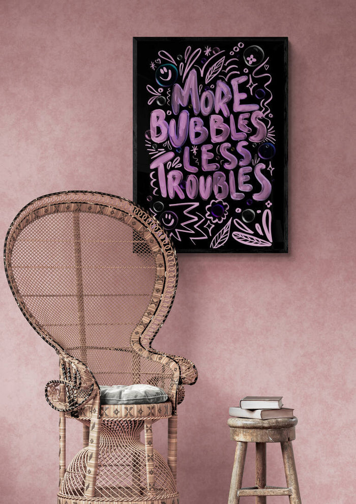 Framed "More Bubbles Less Troubles" Baroo Bloom print on pink wall above wicker chair and small stool with books.