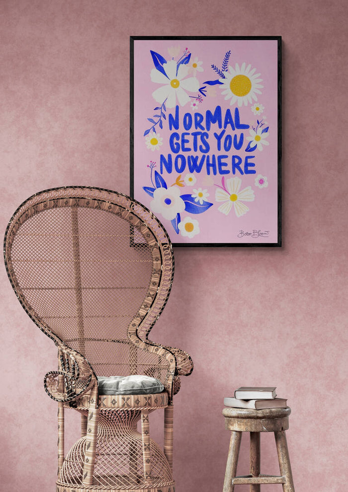 Framed "Normal Gets You Nowhere" wall art with floral design displayed above a wicker chair and stool.