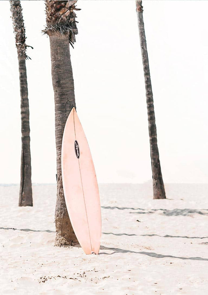 A pink surfboard rests against a palm tree on a sandy beach, with clear sky in the background.