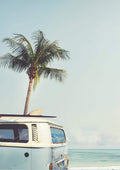 Vintage van with surfboard parked on a beach by the ocean, palm tree in background under clear sky.