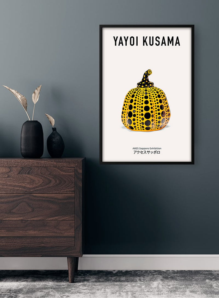 Framed Yayoi Kusama poster featuring a yellow pumpkin with black dots, displayed above a wooden dresser.